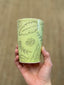 Hand holding a handmade ceramic green cup with leaf patterns and 'The Honey Truck' branding against a beige background