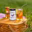 A wooden picnic basket sits on grass outdoors with a glass of iced tea, a jar of honey, and a can of Beekeeper's Tea (Elderberry) arranged on top.