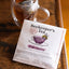 A package of Beekeeper's Tea Elderberry herbal notes tea lies on a dark wooden table next to a clear glass mug filled with amber tea and a tea bag.