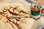 A close-up food scene showing a sliced rustic pastry topped with peaches, and crumbled cheese, served on parchment paper next to an open jar of truffle and rosemary infused honey with a wooden spoon.